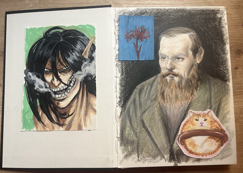 A sketchbook open flat on a wooden table. The left page is a gouache painting of a snarling titan's head against a green background. The right page is a charcoal and colored-pencil portrait of Dostoyevsky, with a small painting of a red spider lily above it and a cut-out of a ginger cat sitting in a bowl.